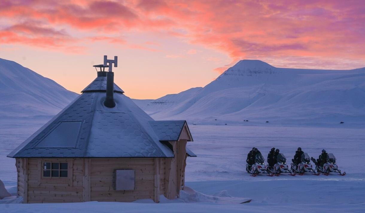 Villmarkshytte og snøskutere i snødekt landskap ved solnedgang.
