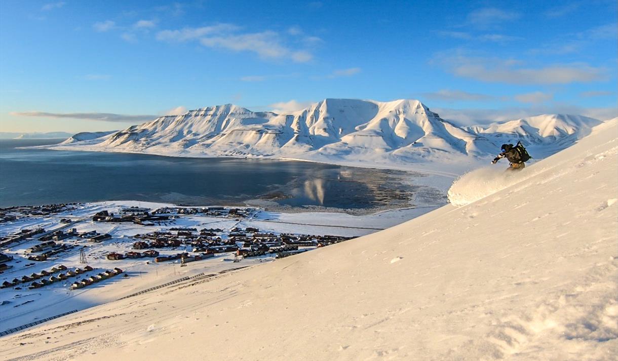 A person skiing down a mountain with Longyearbyen and a mountainous landscape in the background