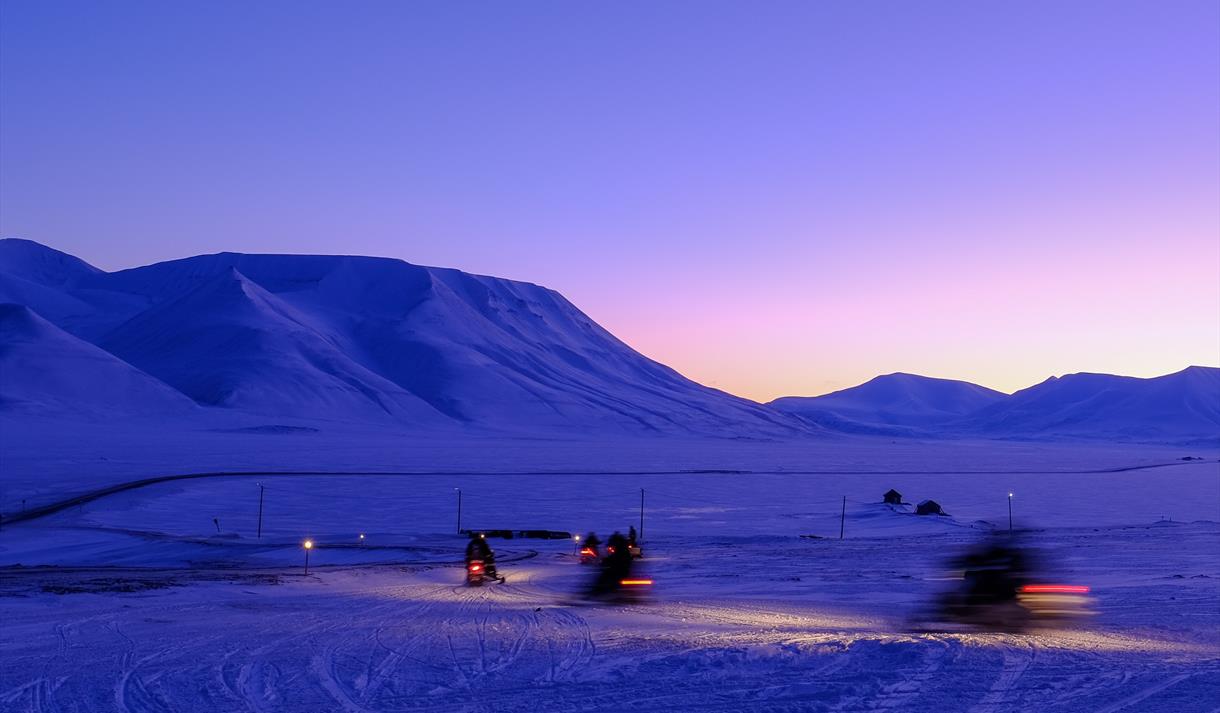 Persons on snowmobiles heading out on a trip with a mountainous landscape covered in blue light in the background