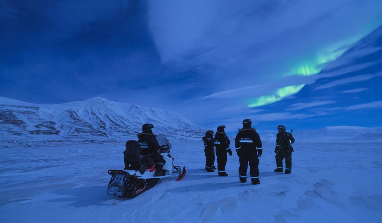 A group of people standing outside with a snowmobile. On the sky its birghr northern lights.