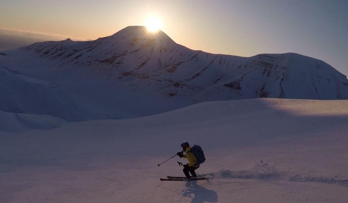 A person skiing down a mountain with powdersnow. Sunset, clear sky and a mountain in the background.