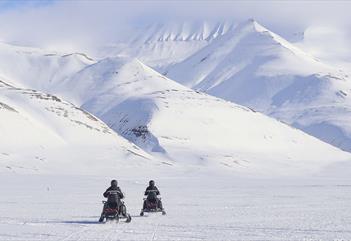 Two persons on electric snowmobiles in the foreground driving across a frozen snowy plain towards snow-covered mountains in the background