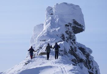Three persons on skis on top of a snowy ridge in the foreground, with the rock formation known as Trollsteinen covered in snow and towering above them