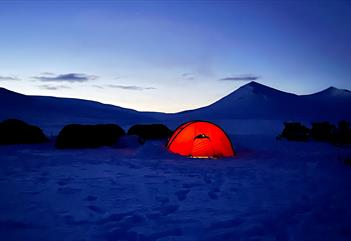 The photo is taken in winter at dusk and shows four tents and some snowmobiles. One of the tents is lit from the inside, glowing red. In the backgroun