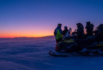 En gruppe med gjester og guide som står bak en snøscooter og ser utover et landskap med blå og oransje skumringsfarger på horisonten