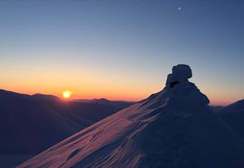 A snow-covered mountain top in the foreground with a low sun and clear skies on the horizon above other mountain tops in the background