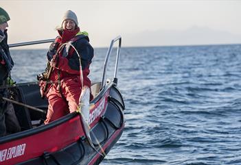 En guide som står ved siden av en gjest i en RIB båt hvor gjesten holder en fiskestang med en fisk på kroken som henger i lufta ved båten