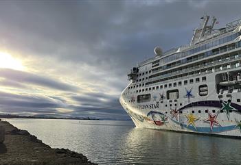 A cruise ship in port inside of a fjord, with an overcast sky and sunshine behind the clouds in the background