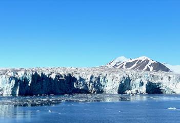 A wide glacier front with a fjord in the foreground and mountains with a clear blue sky in the background