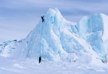 A person climbing a steep ice wall while another stands at the base for safety.