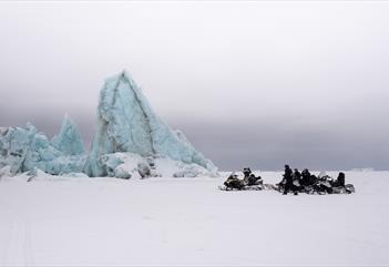 Gjester og en guide på snøscootere som tar en pause ved et isberg frosset inn i sjøis