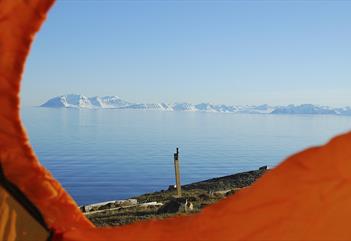 A blue calm fjord with distant snow-covered mountains and a blue sky in the background, seen through the opening of a tent on a hill in the foreground