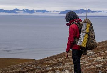 A guide with backpack and rifle on their back, looking out across a fjord in the background
