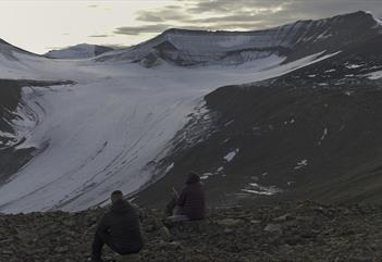 Two guests sitting in a rocky area looking towards the glacier Foxfonna in the background