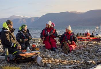 Two guides and two guests sitting on a beach talking with each other, and a RIB boat anchored by the shore of the fjord in the background