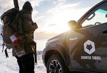 A person carrying a backpack and a rifle on their back standing next to a car, with sunshine, a fjord and a mountain in the background