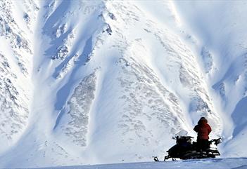 En person som står ved siden av en snøscooter med en massiv snødekt fjellside i bakgrunnen