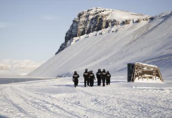 En gruppe gjester i snøscooterutstyr som går mot en bygning i snødekte omgivelser ved Villa Fredheim langs Tempelfjorden, med fjell i bakgrunnen