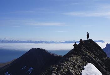 To gjester og en guide på fottur langs en fjellrygg i forgrunne, med en stor skydekt fjord, fjerne fjell og en blå himmel i bakgrunnen