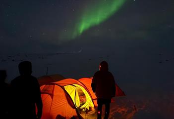 personer står utenfor et opplyst telt på natten på Svalbard å ser på nordlys på himmelen.
