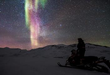 A person stands upright on a parked snowmobile, looking at the clear sky adorned with distinct northern lights and stars.