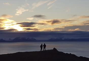 Two persons on top of a mountain in silhouette against a dark fjord and a sunset behind low clouds in the background