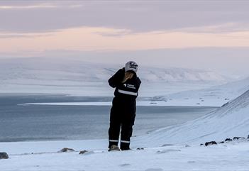 A guest wearing snowmobile equipment standing on a snowy hill scouting out towards a fjord and snowy landscapes in the background