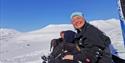 A group of people stopping to take a break to enjoy the sun, with snow-covered mountains in the background. The person in the foreground has a big smi