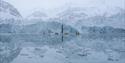 Four kayaks with persons paddling in between floating chunks of ice with a large glacier front in the background