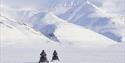 Two persons on electric snowmobiles in the foreground driving across a frozen snowy plain towards snow-covered mountains in the background