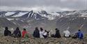 A group of guests and a dog sitting in a line in a rocky field in the foreground, with a valley and mountainous landscapes in the background