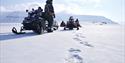 Polar bear tracks on a snowy plain in the foreground, with a guide and several snowmobiles in the background