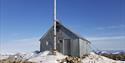 En hytte med metallvegger på toppen av et fjell, omringet av snø og steiner med en blå himmel i bakgrunnen