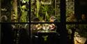 Gjester i Vinterhagen Restaurant som spiser middag med grønne planter i bakgrunnen, sett utenfra gjennom vinduer i restauranten