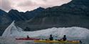 A guide and two guests paddling in kayaks with a floating iceberg and mountain formations in the background