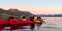 A group of guests in kayaks on a calm fjord with mountains in the background