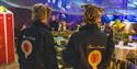 Two persons in overalls with the logo of Mine 3 on their backs. In the background there are restaurant tables and guests.