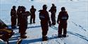 A group of people are standing in a circle in the winter landscape next to a snowmobile. Mountains in the background. Sunny weather.