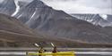 To gjester i en dobbelkajakk som padler over en fjord med fjell i bakgrunnen