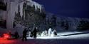 A group of people are standing and walking around some snowmobiles in the dark rigtht below a cliff with frozen ice hanging down from it.
