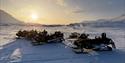 Fire snøscootere som står på snø i et vinterlandskap med solen på hummelen i bakgrunnen.