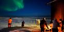 Persons standing next to a bonfire and enjoying the northern lights in the background