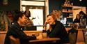 Two guests sitting at a table drinking coffee together, and employees in the background behind the café counter.