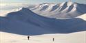 Two persons skiing up a mountain with a long valley and a mountainous landscape in the background