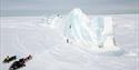 A person climbing a tall ice wall with snowmobiles parked nearby.