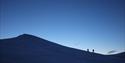 Two persons in silhouette hiking up a snow-covered mountain top, with a clear dark blue sky in the background