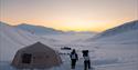 A tent set up in a snow-covered valley with the sunset in the background.