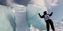 A person stands with his hands in the air in front of a mountain of blue ice.