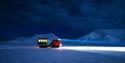 A snowcat in dark snowy surroundings with powerful lights on its front and mountains in the background