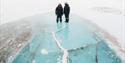 Two persons dressed in snowmobiles suit, standing on blue ice.